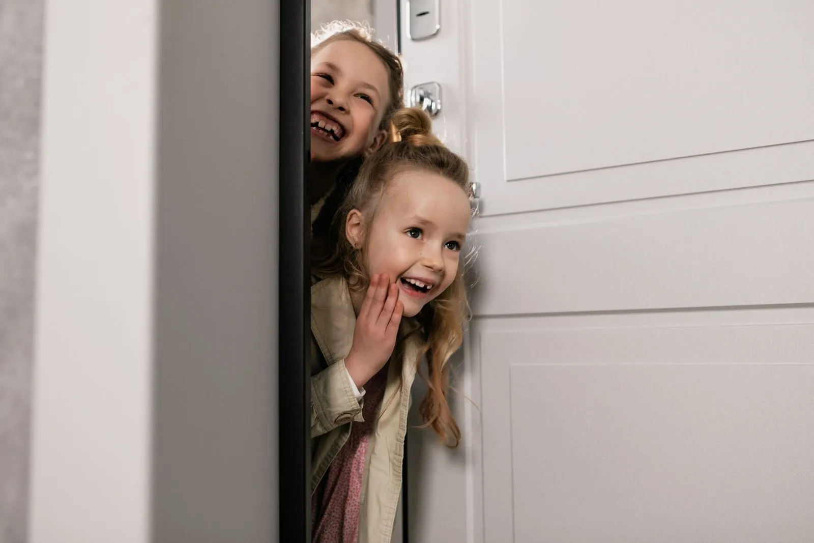 Two happy sisters peeking playfully through a door, smiling indoors.